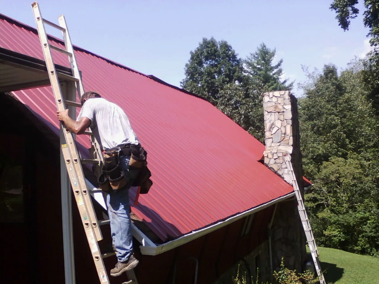 Roofer on a ladder beside a red metal roof and stone chimney in Keyes